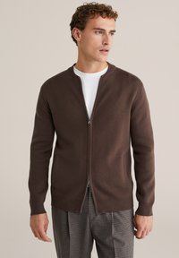 Barna cipzáras pulóvert viselő férfi fehér ing és kockás nadrág fölött, jobbra néz egy egyszínű bézs háttér előtt.