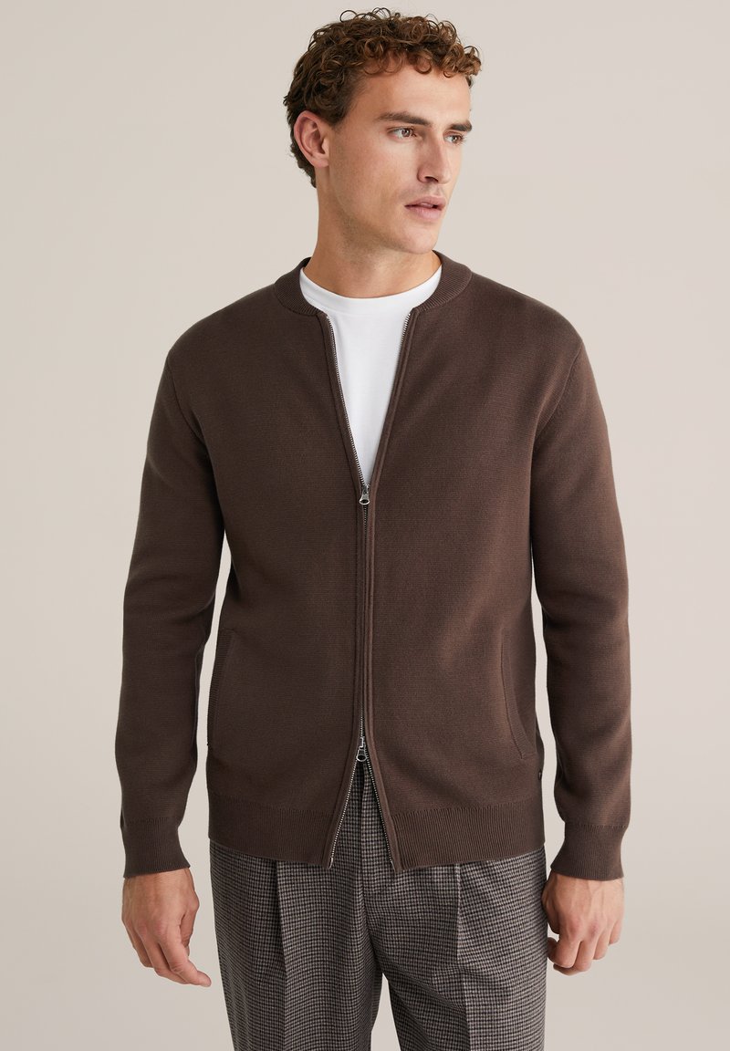 Barna cipzáras pulóvert viselő férfi fehér ing és kockás nadrág fölött, jobbra néz egy egyszínű bézs háttér előtt.