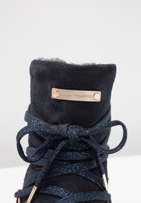 Tmavě modré semišové kotníkové boty s měkkou podšívkou. Mají kovovou destičku s logem a třpytivé tkaničky pro zvýšení detailu.