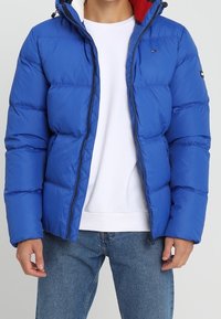 Blauwe pufferjas met een ritssluiting aan de voorkant, voorzien van een capuchon, gequiltde textuur en contrasterende rode voering. Gecombineerd met een wit shirt en blauwe jeans.