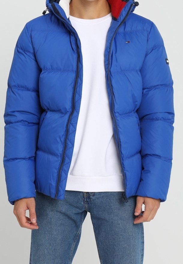 Blauwe pufferjas met een ritssluiting aan de voorkant, voorzien van een capuchon, gequiltde textuur en contrasterende rode voering. Gecombineerd met een wit shirt en blauwe jeans.