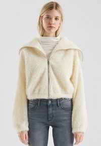 Giacca zip-up di colore crema, morbida e pelosa, con colletto alto e maniche ampie. Indossata su un dolcevita bianco e abbinata a jeans grigio scuro.