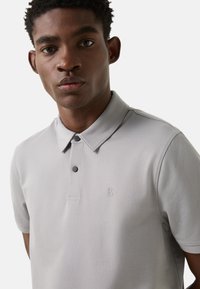 Light grey polo shirt featuring a structured collar, two black buttons, and a subtle logo on the chest. Smooth fabric with a slight texture.
