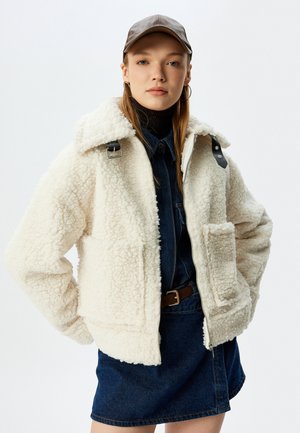 Crème, fluffige fleecejas met een knoopkraag, zijzakken en donkere leren accenten. Draag over een denim jurk met een riem.