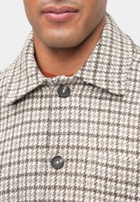 Detailný záber na osobu v hnedo-bielej kárované kabáte s čiernymi gombíkmi a golierom, viditeľné sú pery a brada.