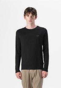 Schwarzes Langarmshirt mit Rundhalsausschnitt und einem dezenten JOOP!-Logo auf der Brust, kombiniert mit hellbeigen Hosen. Weiche Stofftextur.