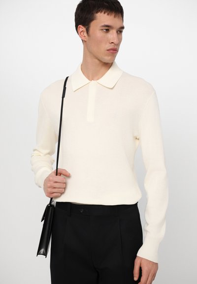 Pólo de malha na cor creme, com mangas longas, apresentando uma pala com botões e uma textura suave. Combinado com calças pretas e uma bolsa preta slim.