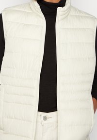 Gepolsterte ärmellose Weste in Cremefarbe mit hohem Kragen, Reißverschlusstasche und diagonal gesteppter Textur. Über einem schwarzen Oberteil getragen.