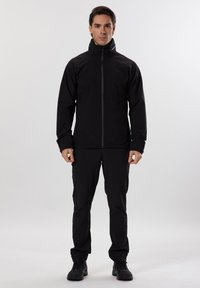 Černá voděodolná bunda s vysokým límcem a předním zipem, doplněná o odpovídající černé kalhoty a černé boty, stojící na neutrálním pozadí.