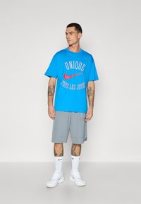 Mėlynas medvilninis marškinėlis su raudonu "Nike" logotipu ir tekstu, derinamas su pilkomis šortais. Outfitą užbaigia baltos sportinės bateliai ir trumpas kojines.
