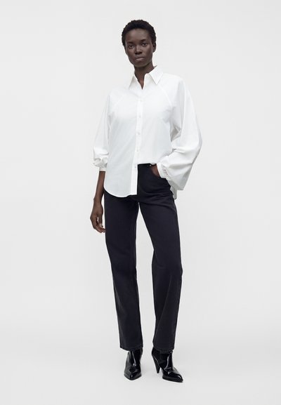 Femme portant une chemise blanche à manches bouffantes, un pantalon noir droit, et des bottines noires à talons, debout sur un fond blanc.
