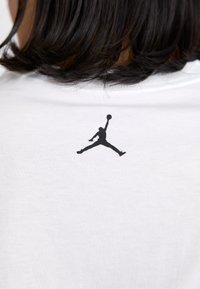 Hvid t-shirt med et sort Jumpman-logo trykt på den øverste ryg. Stoffet ser blødt ud med en glat tekstur og ribbet halsudskæring.