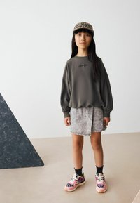Sweatshirt gris avec manches bouffantes, jupe en denim et baskets colorées avec motif léopard. L'arrière-plan présente un triangle texturé sombre.