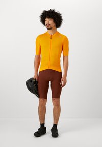 Maillot de cyclisme orange à manches courtes avec une fermeture éclair à l'avant et un tissu texturé, associé à un short rembourré marron ; tenant un casque noir.