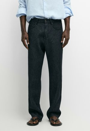 Homme portant un jean noir ample, une chemise bleu clair boutonnée avec les manches retroussées et des sandales noires, debout devant un fond blanc uni.