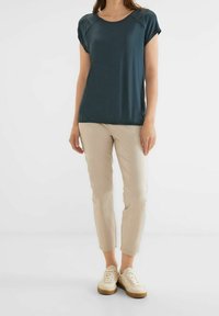 Tmavě tyrkysový top s krátkým rukávem a kulatým výstřihem, zdobený dekorativním prošíváním. Slaďený se světle béžovými slim-fit kalhotami a bílými teniskami.