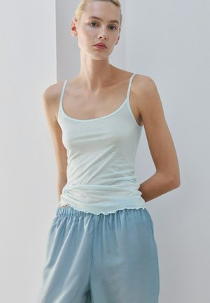 Donna che indossa una canotta azzurra con spalline sottili e pantaloni blu larghi, in piedi davanti a un muro grigio uniforme con le braccia dietro la schiena.