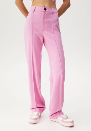 Persoon draagt een roze broek met hoge taille en wijde pijpen, gecombineerd met witte en roze sneakers, staand tegen een effen witte achtergrond.