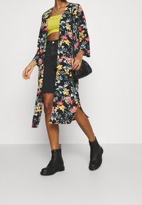 Woman wearing a black floral long cardigan, lime green crop top, black denim skirt, black boots, and holding a black handbag while walking.