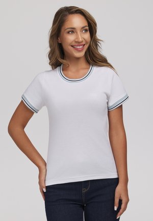 Uśmiechnięta kobieta o długich włosach, ubrana w białą koszulę z krótkim rękawem ozdobioną paskami oraz ciemne dżinsy, stojąca na jednolitym tle.