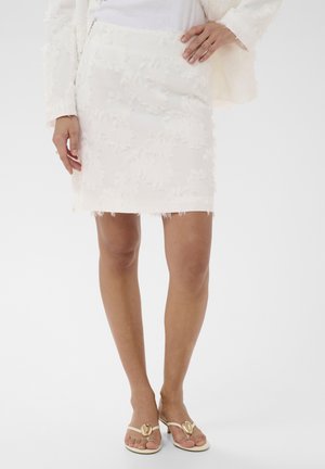 Femme portant une jupe blanche texturée et des sandales blanches à talons ornées de décorations en forme de cœur dorées, debout devant un fond blanc uni.