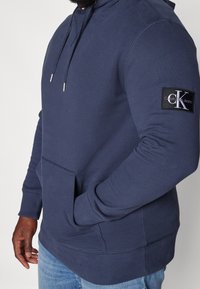 Felpa blu navy realizzata in morbido cotone misto, con tasca frontale, cappuccio con coulisse regolabile e un'etichetta con logo sulla manica.