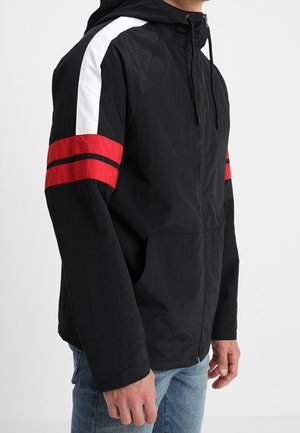 Homme portant une veste à capuche noire avec des bandes blanches et rouges sur les manches et un jean bleu, debout devant un fond clair uni.