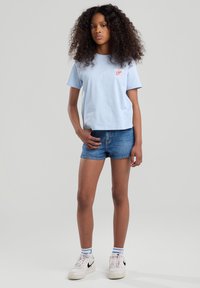T-shirt di cotone azzurro chiaro con maniche corte e logo rosso, abbinato a pantaloni corti in denim. Il modello indossa sneaker bianche e calze a righe.