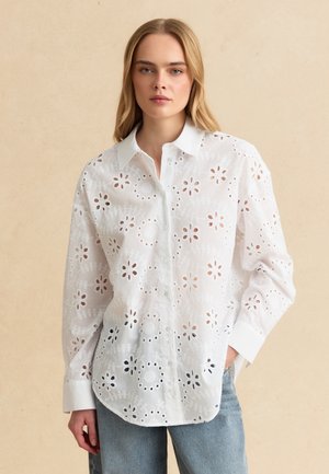 Jeune femme portant un chemisier blanc à manches longues ajouré avec des découpes florales et un jean bleu clair, debout devant un fond beige.