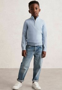 Világoskék zipzáras pulóver, amelyet felgöngyölt, laza szabású farmernadrággal és fehér sportcipővel visel, semleges háttér előtt állva.