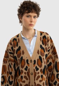 Pletený cardigan s výstřihem do V, hnědým, černým a oranžovým leopardím vzorem na béžovém pozadí, se čtyřmi knoflíky a spuštěnými rameny.