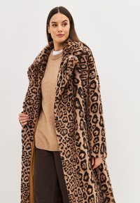 Abrigo de piel de leopardo con un cuello ancho, con patrones en marrón y negro. Llevado sobre un suéter beige, tiene un diseño largo y fluido.