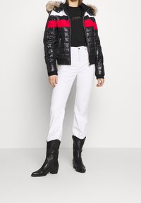 Vadderad svart jacka med röda och vita horisontella ränder, huva med päls, svart tröja, vita jeans och svarta ankelstövlar.