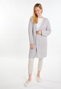 Cardigan tricotat de culoare gri deschis, cu mâneci lungi, deschis în față și două buzunare frontale, purtat peste un top alb și pantaloni bej. Textură moale.
