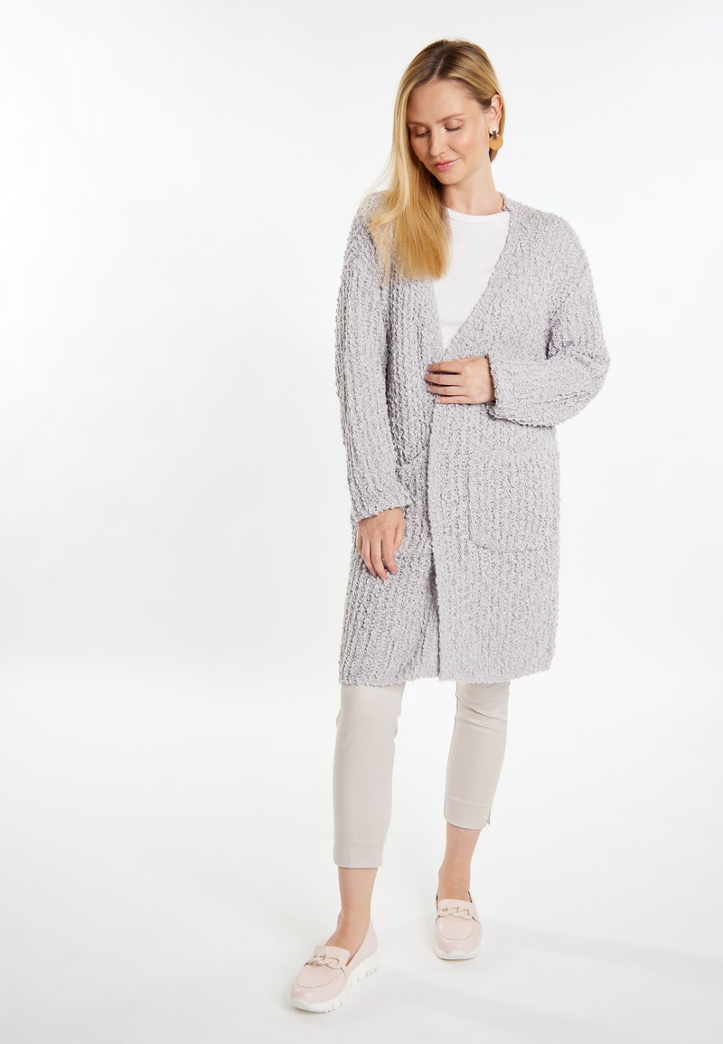 Cardigan tricotat de culoare gri deschis, cu mâneci lungi, deschis în față și două buzunare frontale, purtat peste un top alb și pantaloni bej. Textură moale.
