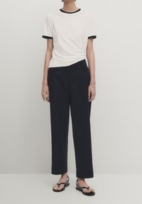 Biała koszulka z czarnym wykończeniem, w połączeniu z ciemnoniebieskimi szerokimi spodniami. Sandały z cienkimi paskami dopełniają stylizację, podkreślając swobodny wygląd.