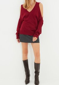 Bordowy oversize sweter z dekoltem w serek i odkrytymi ramionami, zestawiony z ciemnoszarą mini spódnicą i brązowymi kozakami za kolano.