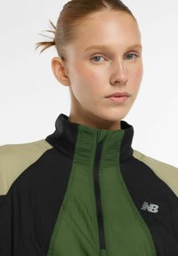 Jonge vrouw met opgestoken haar, draagt een groen en zwart New Balance vest met rits, neutrale uitstraling, lichte achtergrond.