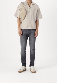 Uomo che indossa una camicia button-up a maniche corte color beige chiaro, jeans slim-fit grigi, sneakers bianche e una borsa a tracolla beige su uno sfondo bianco.