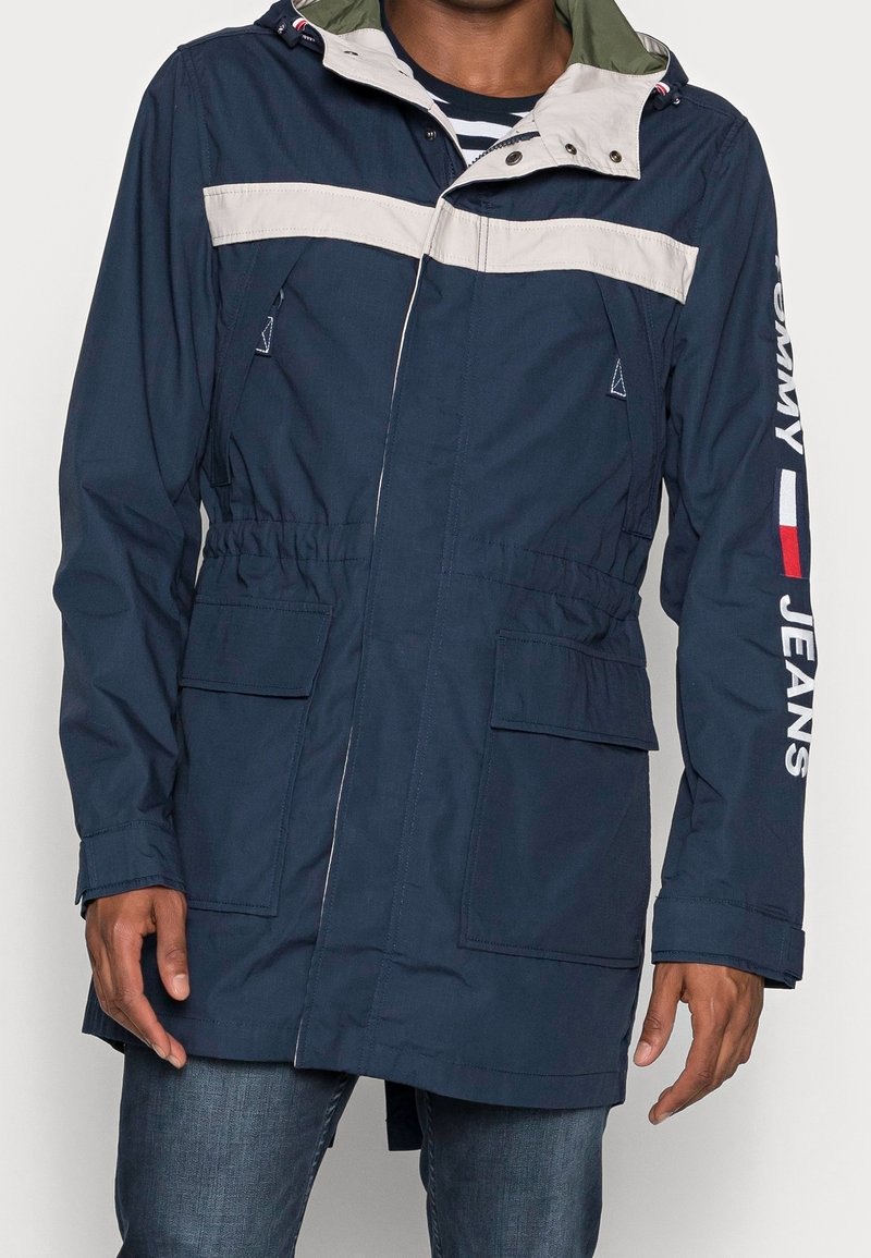 Námořnická modrá voděodolná parka s kontrastním bílým pruhem na hrudi, nastavitelnou kapucí, předními kapsami a brandingem "Tommy Jeans" na rukávu.