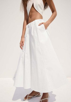 Femme portant une jupe blanche taille haute en ligne A avec des poches et des sandales blanches à lanières, debout devant un fond clair uni.
