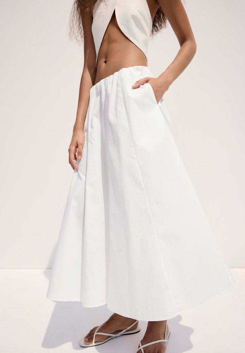 Femme portant une jupe blanche taille haute en ligne A avec des poches et des sandales blanches à lanières, debout devant un fond clair uni.