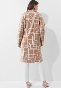 Bruine lang coat met een patroon van witte geometrische ontwerpen, met een relaxte fit en lange mouwen, gecombineerd met witte broek en lichtgekleurde schoenen.