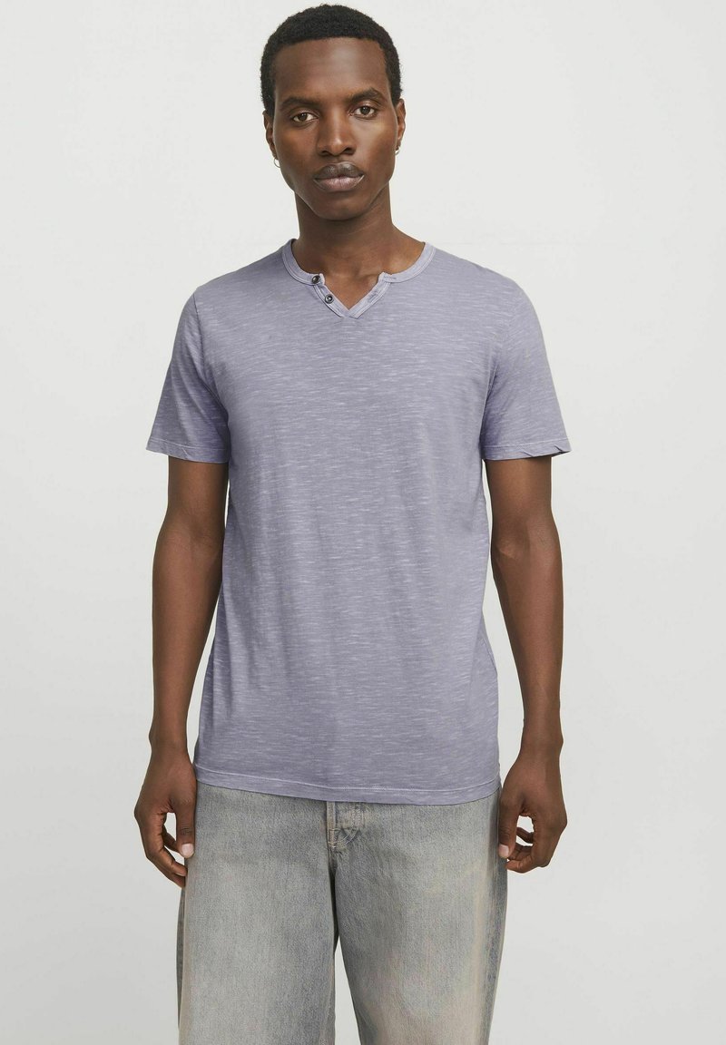 Camiseta corta de manga corta en un suave color lila con cuello en V y dos botones. Hecha de una tela suave y texturizada. Llevada con pantalones cortos de mezclilla claros.