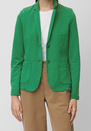 Mujer con una chaqueta blazer verde brillante sobre una camiseta blanca y pantalones marrones, de pie con los brazos relajados a los lados.