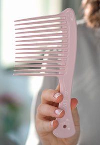 Różowy plastikowy grzebień z długimi, prostymi zębami, lekko zakrzywioną rączką i okrągłym otworem na końcu do zawieszania. Czysty design i gładka tekstura.