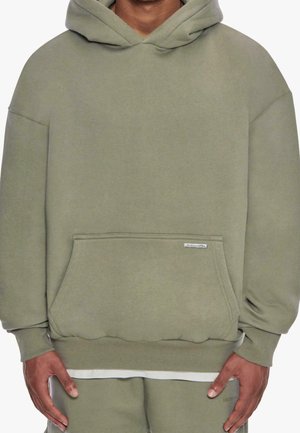 Mann trägt olivgrünen Hoodie mit Fronttasche und passende Hose, steht vor weißem Hintergrund.