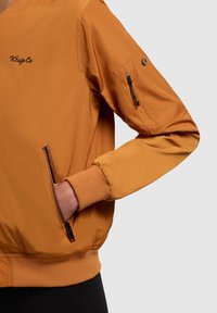Oranje bomberjack met een ritssluiting voorvak, ribgebreide manchetten en een geborduurd logo op de borst, met een gladde textuur.