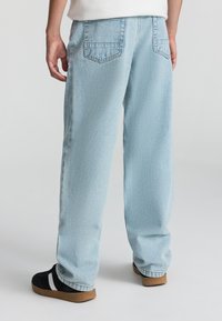 Jeans in denim azzurro chiaro con vestibilità rilassata, dotati di tasche posteriori e una superficie leggermente strutturata, abbinati a scarpe nere.