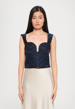 Navy cropped top with pleated texture, featuring a deep V-neck and wide straps. Worn with a satin beige skirt.